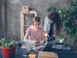 Young designer is drawing images in notebook sitting at table while female photographer is coming to her with camera. Women start discussing new project and watching photos on screen.