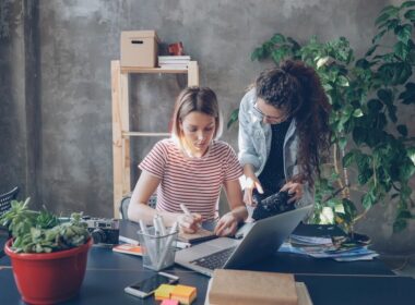 Young designer is drawing images in notebook sitting at table while female photographer is coming to her with camera. Women start discussing new project and watching photos on screen.