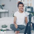 Female blogger is recording video for videolog looking at camera, smiling and talking emotionally sitting on bedroom floor at home. People, technology and social media concept.