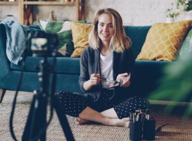 Make-up artist and blogger is recording video about face contouring for her vlog using professional camera. Young woman is holding brush and cosmetics kit and speaking.