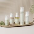 A beautifully arranged display of elegant skincare products on a marble tray against a soft, beige backdrop. The collection includes various bottles and jars, showcasing a range of serums, creams, and SPF protection. The minimalist design and soft pastel colors create a clean, luxurious aesthetic. A delicate floral arrangement in a textured vase adds a natural touch to the composition, enhancing the serene and sophisticated vibe.