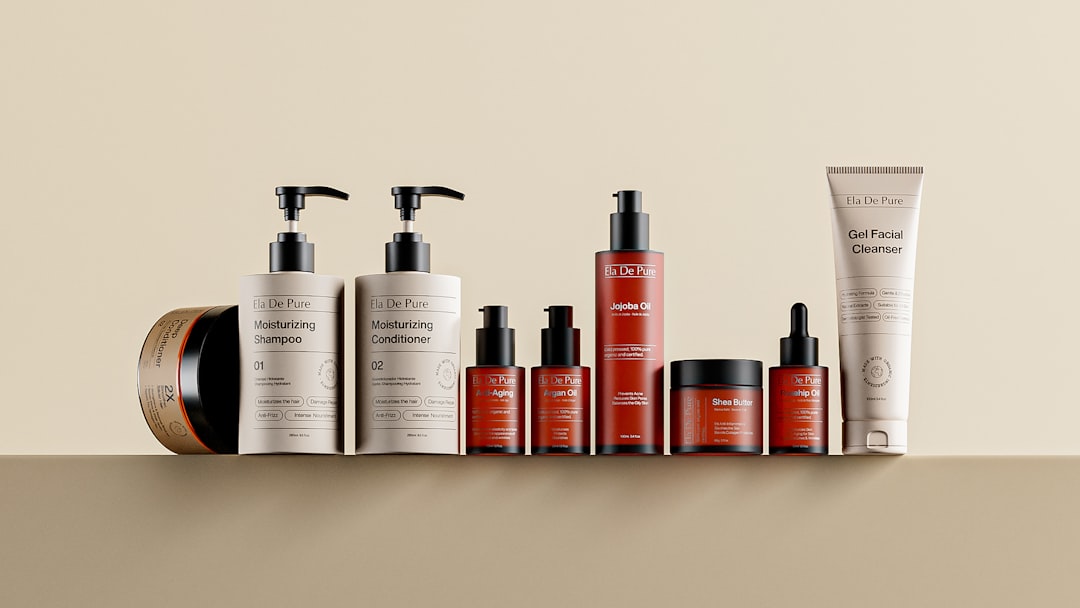 A clean, minimal studio composition featuring a premium lineup of Ela De Pure skincare and haircare products arranged in a balanced row against a warm beige background. The collection includes moisturizing shampoo and conditioner bottles with matte pumps, treatment oils in amber dropper bottles, a deep conditioning hair masque jar, a jojoba oil bottle, a shea butter treatment, and a gel facial cleanser tube. Soft lighting highlights the contrast between matte packaging and glossy elements, giving the lineup a refined, professional beauty brand aesthetic that emphasizes simplicity